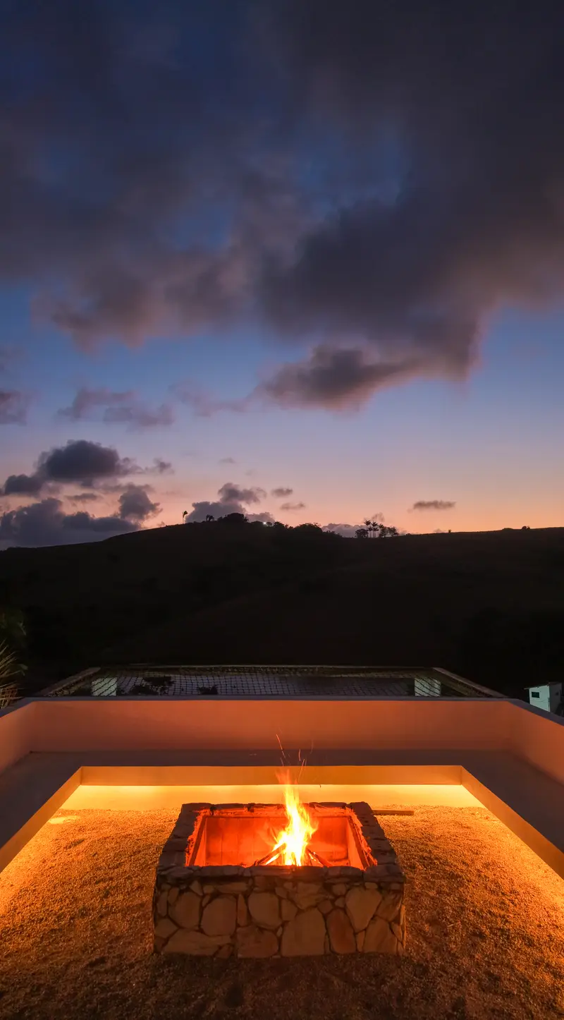 Fogueira acesa em um recanto de pedra ao anoitecer, com as colinas e o céu entre azul e rosa ao fundo