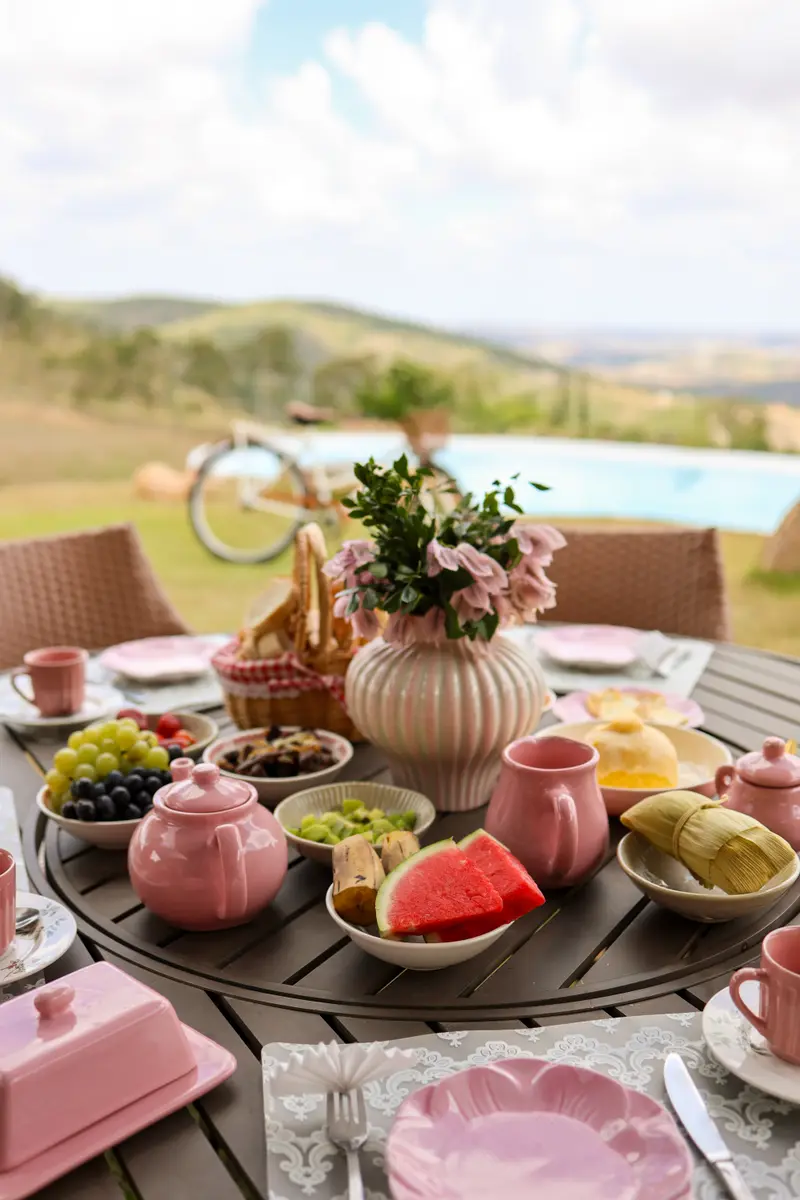 Mesa redonda de café da manhã com louça rosa e frutas, com a piscina ao fundo